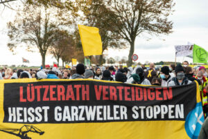 A demonstration arm/Finger moves through Lützerath. In the foreground is a banner with the statement "Defending Lützerath means stopping Garzweiler".