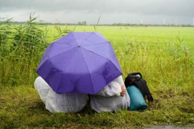 zwei Menschen von hinten in weißen Aktionsanzügen sitzen Arm in Arm unter einem blauen Regenschirm an einem Feldrand.