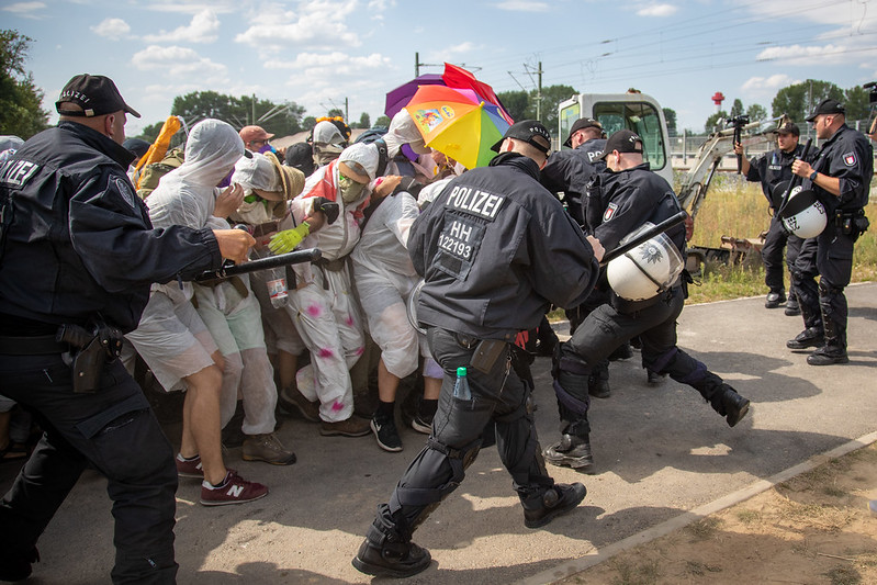 Blockade eines Fingers trifft auf die Polizei, ein Schirm in Regenbogenfarben ist zu sehen.