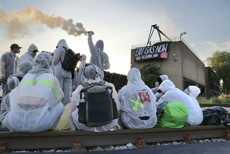 Menschen sitzen auf Schinen. Im Hintergrund ist ein Stapel von Pipelineteilen zu sehen und auf einem großen Banner steht "Exit Gas Now"