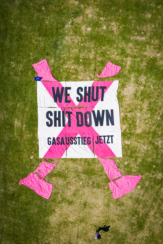 Auf einer Wiese liegt ein großes Banner auf dem steht "We shut shit down. Gasausstieg Jetzt"
