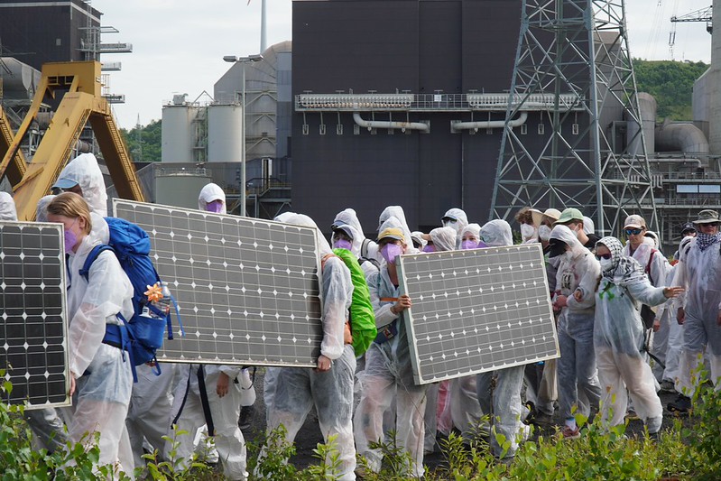 Menschen in weißen Maleranzügen laufen über ein Kraftwerksgelände und tragen Solarpanele